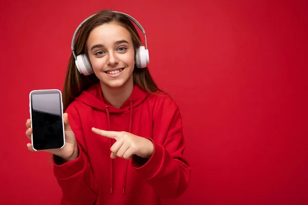 Sonriente hermosa dama morena con capucha roja aislada sobre fondo rojo sosteniendo y mostrando teléfono inteligente con pantalla vacía para recortar usando auriculares inalámbricos blancos escuchando música divertida — Foto de Stock