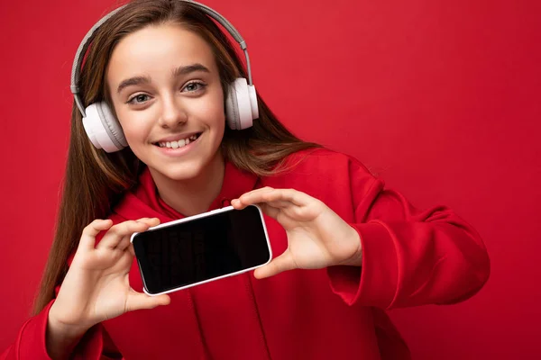 Zbliżenie portret amazibg piękne brunetka dziewczyna w czerwonym kapturze izolowane na czerwonym tle gospodarstwa i pokazując smartfon z pustym wyświetlaczem do cięcia noszenia białe słuchawki bezprzewodowe słuchanie — Zdjęcie stockowe