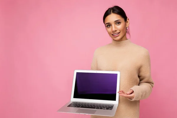 Hermosa mujer joven con cabello oscuro mirando a la cámara que sostiene el ordenador portátil con pantalla de monitor vacía con maqueta y espacio de copia con suéter de color claro aislado en el fondo de la pared rosa —  Fotos de Stock