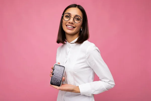 Fénykép bájos mosolygós fiatal barna nő visel fehér blúz és optikai szemüveg áll elszigetelt felett rózsaszín háttér mutatja mobiltelefon üres képernyőn mockup nézi a kamerát — Stock Fotó