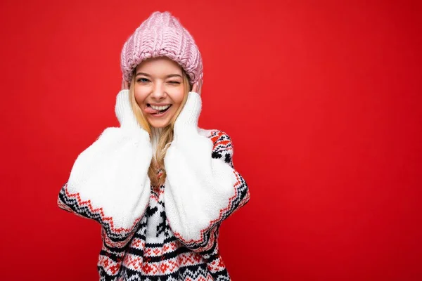 Foto de hermosa feliz divertida alegre joven rubia aislada sobre la pared de fondo rojo con suéter cálido invierno y sombrero rosa mirando a la cámara guiñando y mostrando la lengua — Foto de Stock