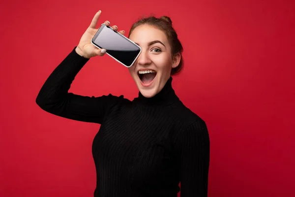 검정 스웨터를 입고 빨간 배경에 고립된 채서 있는 매력적 인 젊은 갈색 머리 여자의 사진은 카메라를 보기 위해 빈 화면이 달린 휴대 전화를 보여 주고 있다. — 스톡 사진