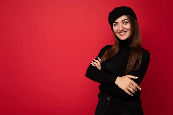 Fotografie krásné šťastné usmívající se mladé brunetky žena izolované přes červené pozadí zeď na sobě černý svetr a černý klobouk při pohledu na kameru — Stock fotografie