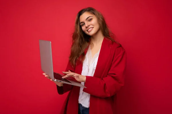 Photo of Beautiful smiling young brunet woman holding netbook computer typing on keyboard looking at camera wearing red cardigan and white t-shirt isolated on red wall background — Stock Photo, Image