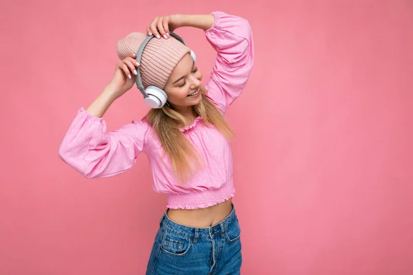 Bella felice sorridente giovane donna bionda indossa camicetta rosa e cappello rosa isolato su parete di sfondo rosa indossando cuffie bluetooth wireless bianche ascoltare musica cool divertirsi e — Foto Stock