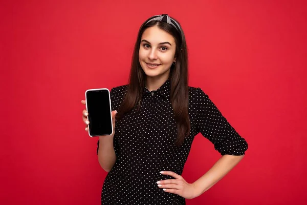 Vrij lachend gelukkig brunette meisje staan geïsoleerd over rode muur dragen casual stijlvolle zwarte kleren tonen mobiele telefoon met leeg scherm voor cutout kijken naar camera — Stockfoto