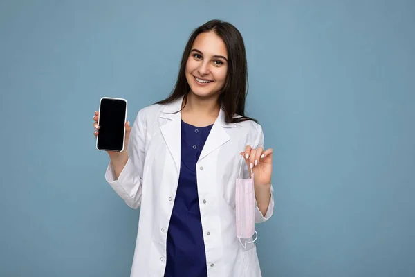 Photo de belle jeune femme brune souriante belle vêtue d'un manteau médical blanc isolé sur fond bleu avec espace de copie tenant smartphone montrant le téléphone à la main avec écran vide — Photo