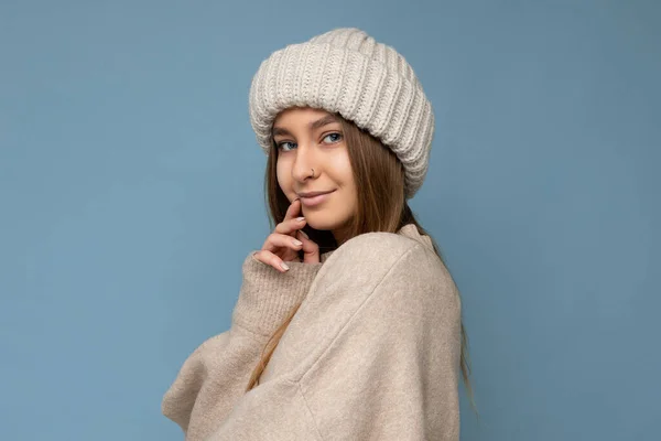 Acogedor lindo disparo de atractivo encantador fascinante bastante feliz joven rubia oscura mujer de pie aislado en la pared de fondo azul con suéter beige cálido y sombrero beige de invierno mirando a la cámara —  Fotos de Stock