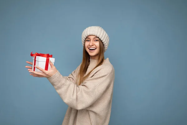 Skott av attraktiva positiva leende ung mörkblond kvinna isolerad över färgglada bakgrundsvägg bär varje trendig outfit hålla nytt år presentförpackning med rött band och ha kul att titta på — Stockfoto