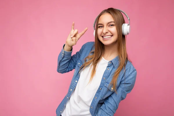 Photo shot of beautiful positive smiling young blonde woman wearing white wireless bluetooth headsets listening to cool music and having fun looking at camera and showing finger up — Stock Photo, Image