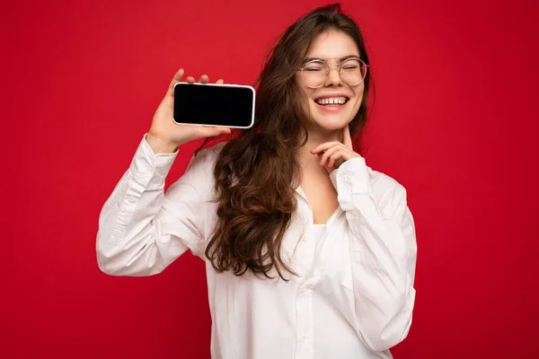 Sorrindo atraente jovem morena vestindo camisa branca e óculos ópticos isolados sobre fundo vermelho segurando na mão e mostrando telefone celular com tela vazia para maquete e se divertindo — Fotografia de Stock