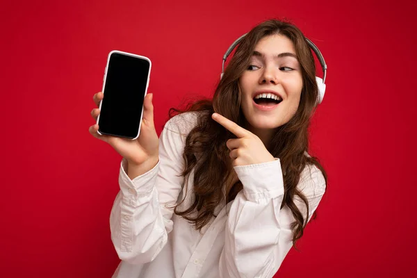 매력적 인 섹시하고 긍정적 인 젊은 갈색 곱슬머리의 여성붉은 배경 벽에 고립된 흰색 셔츠를 입고 흰색 헤드폰을 낀 빈 디스플레이가 달린 휴대 전화를 보여 주고 있다. — 스톡 사진