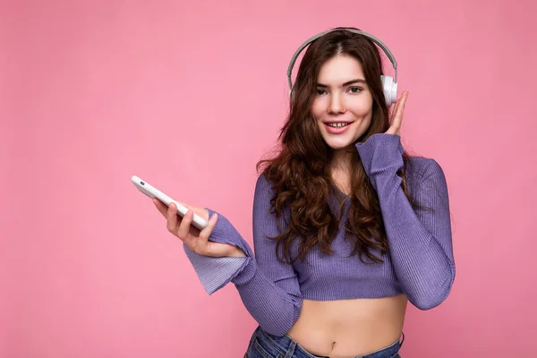 Foto de hermosa mujer joven alegre sonriente con ropa casual elegante aislada sobre la pared de fondo sosteniendo y utilizando el teléfono móvil con auriculares bluetooth blancos escuchando música y — Foto de Stock