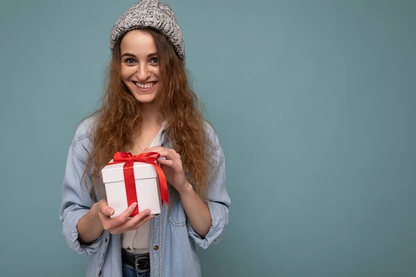 Piękne szczęśliwy młody ciemny blondynka kobieta izolowane nad kolorowe tło ściany noszenie stylowe ubrania casual trzymając pudełko prezentów i patrząc na aparat — Zdjęcie stockowe