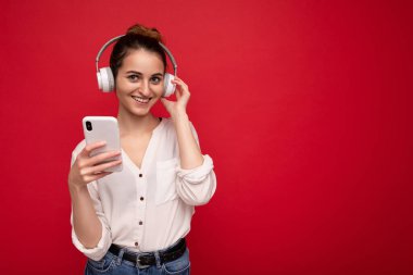 Renkli arka plan duvarından izole edilmiş, beyaz bluetooth kablosuz kulaklıklar takan ve müzik dinleyen güzel, neşeli, güler yüzlü, gündelik kıyafetler giyen genç bir kadının fotoğrafı.
