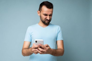 Memnun olmayan, sakallı, mavi tişörtlü, mavi arka planda izole edilmiş ve internetten cep telefonu iletişimi kullanan üzgün ve yakışıklı bir adamın fotoğrafı.