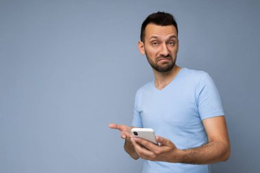 Düşünceli yakışıklı genç adam arka plan duvarından izole edilmiş günlük giysiler giyiyor ve cep telefonu yazıları yazıyor kameraya bakıyor ve düşünüyor.