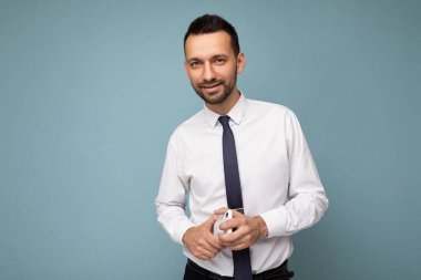Yakışıklı, esmer, sakallı, beyaz gömlekli ve kravatlı bir adamın fotoğrafı. Mavi arka planda izole edilmiş, el telefonunda boş bir alanda kameraya bakıyor.