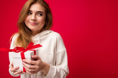 Çekici, pozitif gülümseyen genç, sarışın bir kadın. Renkli arka plan duvarından izole edilmiş, günlük moda kıyafetler giymiş, elinde hediye kutusuyla yan tarafa bakıyor.