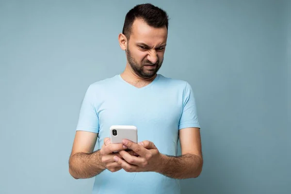 Photo of dissatisfied sad handsome young man with beard wearing everyday blue t-shirt isolated over blue background holding and using mobile phone communication online on the internet looking at — Stock Photo, Image