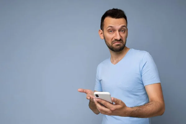 Pensativo joven guapo aislado sobre la pared de fondo usando ropa cotidiana sosteniendo y usando sms de escritura de teléfono móvil mirando a la cámara y pensando —  Fotos de Stock