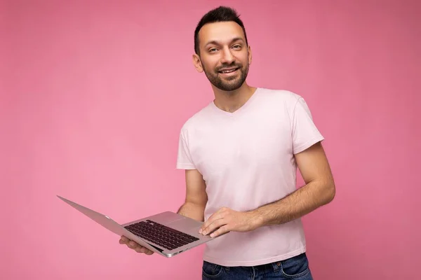 Hombre sonriente guapo sosteniendo ordenador portátil mirando a la cámara en camiseta sobre fondo rosa aislado — Foto de Stock