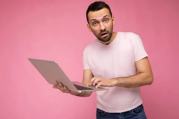 Bonito homem surpreso e surpreso segurando computador portátil olhando para a câmera em t-shirt no fundo rosa isolado — Fotografia de Stock