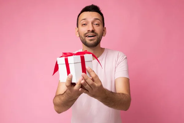 Guapo positivo increíble morena joven hombre sin afeitar con barba aislada sobre la pared de fondo rosa con camiseta rosa sosteniendo caja de regalo blanca con cinta roja y mirando a la cámara —  Fotos de Stock