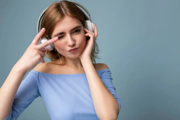 Photo of charming positive sexy young blonde woman wearing blue crop top isolated on blue background wearing white wireless bluetooth headphones listening to cool music and having fun showing peace — Stock Photo, Image
