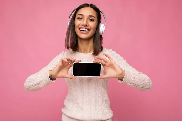 Belle jeune femme souriante heureuse portant une tenue décontractée élégante isolée sur le mur de fond tenant et montrant le téléphone portable avec écran vide pour maquette portant des écouteurs bluetooth blancs — Photo