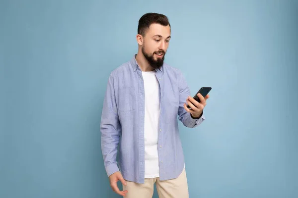 Guapo joven morena sin afeitar hombre con barba usando elegante camiseta blanca y camisa azul aislado sobre fondo azul con espacio vacío sosteniendo en la mano y el uso de sms de mensajería telefónica mirando — Foto de Stock