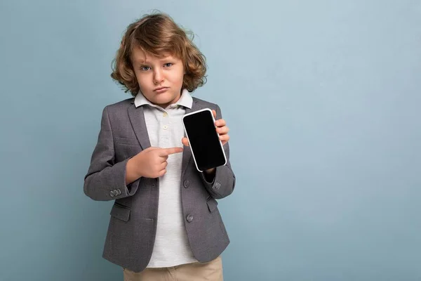 Menino bonito com cabelo encaracolado vestindo terno segurando telefone isolado sobre fundo azul olhando para a câmera e mostrando smartphone com tela de exibição vazia e apontando para a tela — Fotografia de Stock