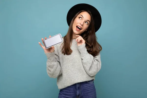 생각있고 착해 보이는 젊고 행복 한 젊은 여성 이 검정 모자와 회색 스웨터를 입고 배경에서 고립된 전화기를 들고 올려다보고 있는 모습 — 스톡 사진