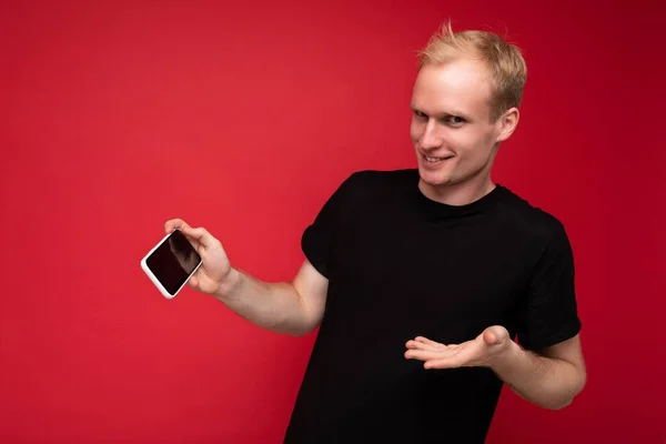 Guapo positivo feliz rubia joven hombre vistiendo camiseta negra de pie aislado sobre fondo rojo con espacio de copia celebración de teléfono inteligente que muestra el teléfono en la mano con pantalla vacía para maqueta —  Fotos de Stock