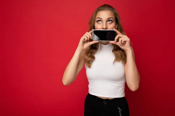 Atraente jovem loira mulher pessoa vestindo branco t-shirt isolado no fundo vermelho com espaço de cópia segurando smartphone mostrando telefone na mão com tela vazia para recorte olhando para cima — Fotografia de Stock