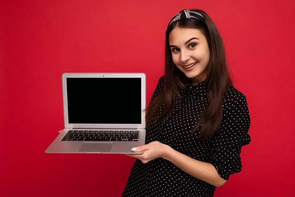 Fénykép gyönyörű mosolygós barna lány visel fekete ruhát gazdaság számítógép laptop nézi kamera elszigetelt színes fal háttér — Stock Fotó