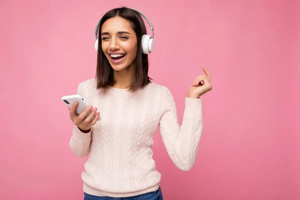 Zdjęcie pięknej, radosnej, uśmiechniętej młodej kobiety w stylowych, luźnych ciuchach odizolowanych nad ścianą tła, trzymającej i używającej telefonu komórkowego w białych słuchawkach bluetooth słuchających muzyki i — Zdjęcie stockowe