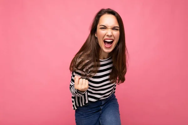 Retrato de joven positivo feliz hermosa morena mujer persona con emociones sinceras usando suéter de rayas casuales aislados sobre fondo rosa con espacio vacío y celebrando ganar gritando sí —  Fotos de Stock