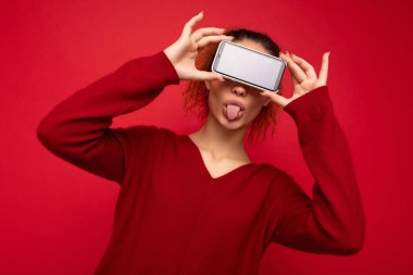 Kırmızı arka plan üzerinde koyu kırmızı süveter giyen mutlu genç bir kadın akıllı telefonu tutuyor ve cep telefonu ekranını boş bir şekilde gösteriyor.