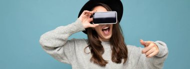 Siyah şapkalı ve gri kazaklı çekici genç bir kadının arka planda izole edilmiş kamerayı işaret ederek parmak kaldırdığı panoramik yakın plan.