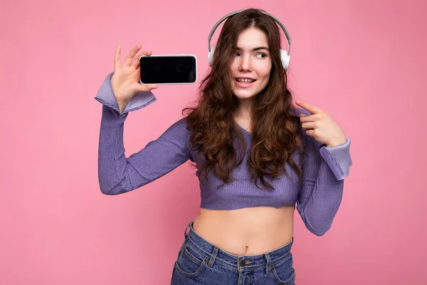 Attractive positive smiling young woman wearing stylish casual outfit isolated on colourful background wall holding and showing mobile phone with empty screen for cutout wearing white bluetooth — Stock Photo, Image