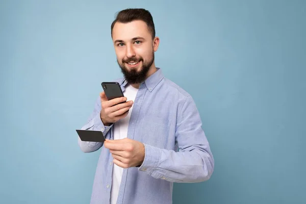 Hombre guapo que usa ropa cotidiana aislada en la pared de fondo sosteniendo y usando el teléfono y la tarjeta de crédito haciendo el pago mirando a la cámara —  Fotos de Stock