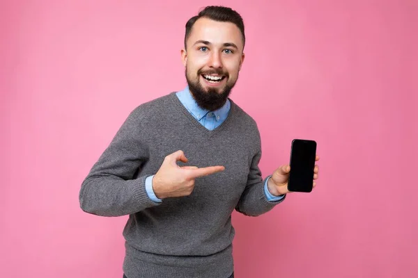 Фото красивого улыбающегося взрослого мужчины, хорошо выглядящего в повседневной одежде, стоящего изолированно на заднем плане с копировальным пространством со смартфоном, показывающим телефон в руке с пустым дисплеем экрана для — стоковое фото