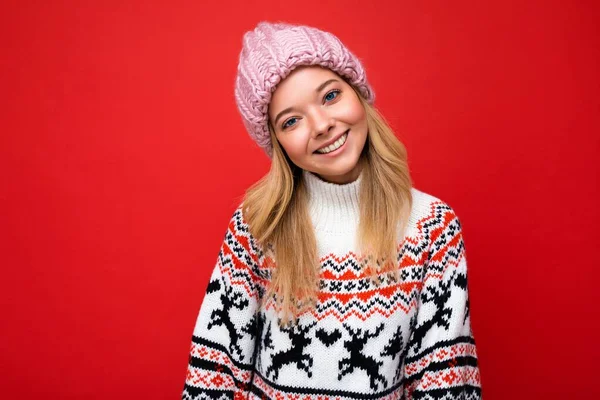 Foto de hermosa mujer rubia sonriente feliz aislada sobre la pared de fondo rojo con suéter de invierno y sombrero rosa de moda mirando a la cámara — Foto de Stock