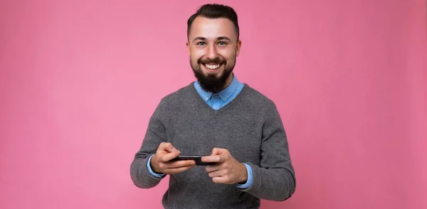 Foto panorâmica de legal boa aparência morena sem barba jovem com barba vestindo camisola cinza e camisa azul isolada sobre a parede de fundo rosa com espaço vazio segurando na mão e usando celular — Fotografia de Stock