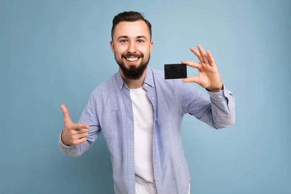 Фото красивой улыбающейся брюнетки бородатого молодого человека в стильной синей рубашке и белой футболке, изолированной на синем фоне, держащей кредитную карту, смотрящей в камеру — стоковое фото