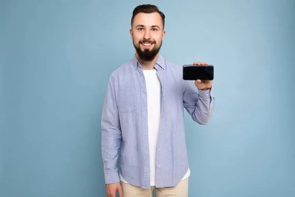 Photo of serious handsome good looking young brunette unshaven man with beard wearing casual white t-shirt and blue shirt poising isolated on blue background with empty space holding in hand mobile — 图库照片