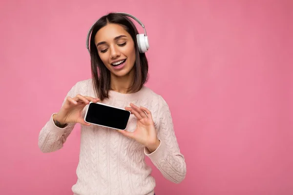 Attractive positive smiling young woman wearing stylish casual outfit isolated on colourful background wall holding and showing mobile phone with empty screen for cutout wearing white bluetooth — Stock Photo, Image