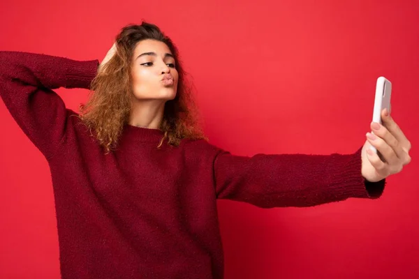 Attractive young blogger woman with curly hair wearing dark red sweater isolated on red background wall holding and using smart phone looking at telephone screen and taking selfie and giving kiss — Stock Photo, Image
