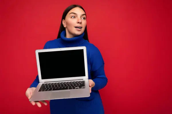 Fascinante sorprendido bastante feliz hermosa joven morena mujer sosteniendo ordenador portátil mirando a un lado usando suéter azul oscuro aislado sobre fondo de pared roja — Foto de Stock
